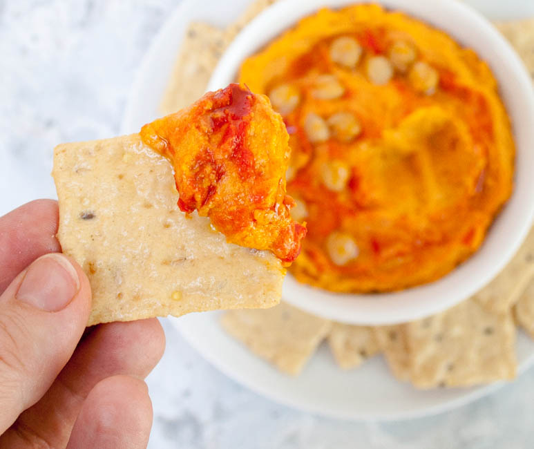 Harissa Hummus in hand with cracker. Bowl of hummus in the background.