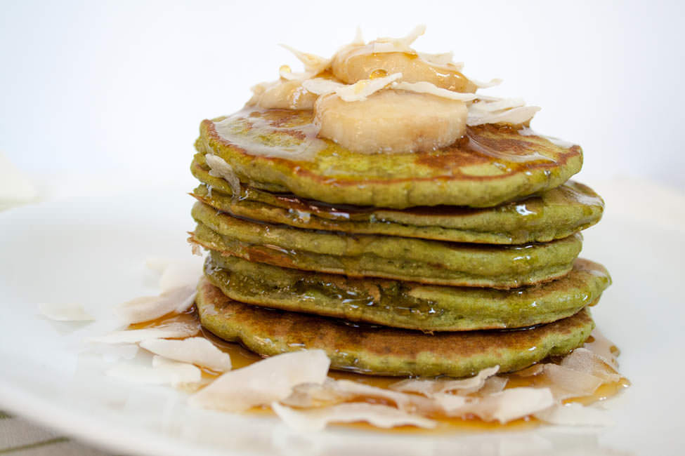 Matcha Banana Pancakes stacked with a drizzle of maple syrup, sliced banana, and coconut flakes.