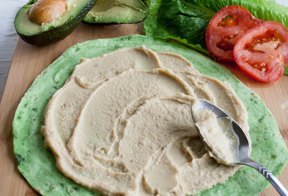 Hummus layered on a tortilla with avocado, lettuce, and tomato in the background for an ALT (Avocado, Lettuce, and Tomato) Wrap.