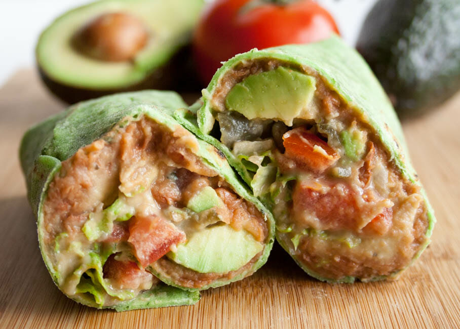 Refried Bean Burrito Recipe close up with avocados and tomatoes in the background.