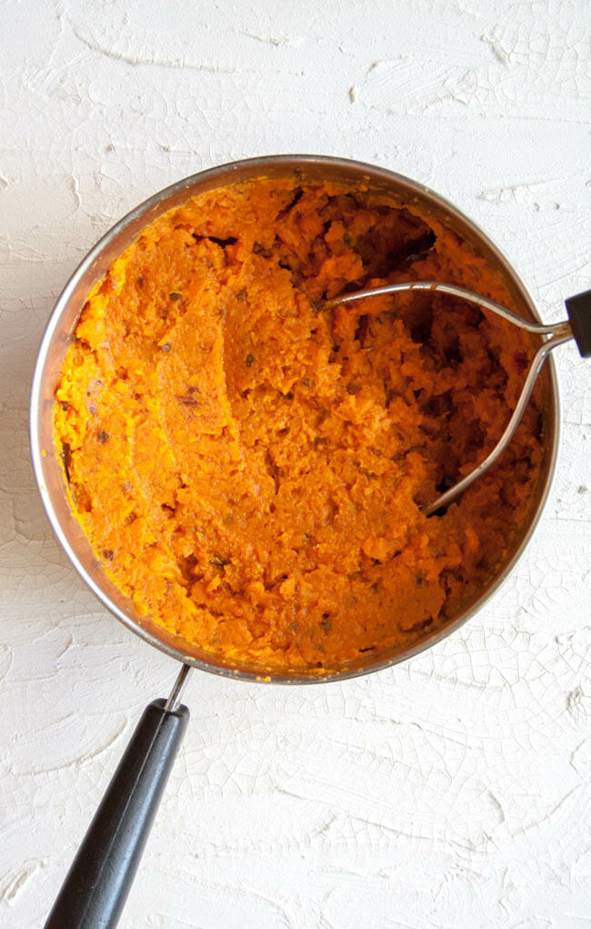 Mashed sweet potatoes in a sauce pan with potato masher.