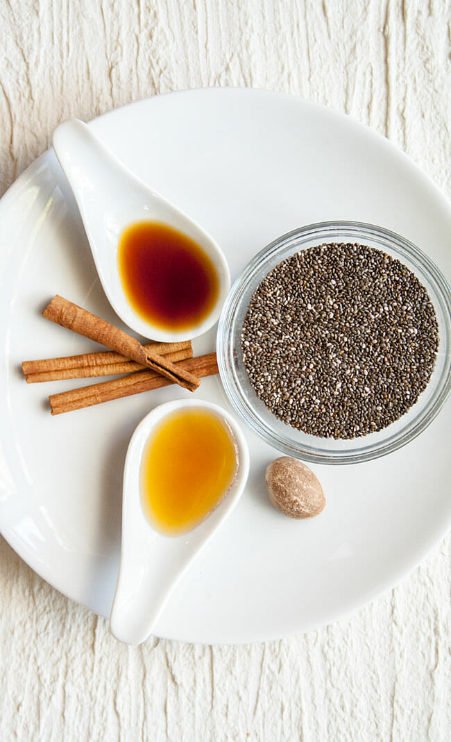 Spoonfuls of maple syrup and vanilla extract with bowl of chia seeds, cinnamon sticks, and whole nutmeg on a plate.