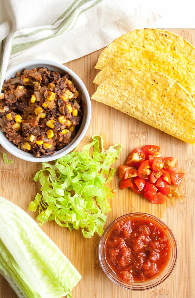 Vegan taco ingredients on a cutting board: black bean and corn mixture, chopped lettuce, tomatoes, salsa, and hard shell tacos.