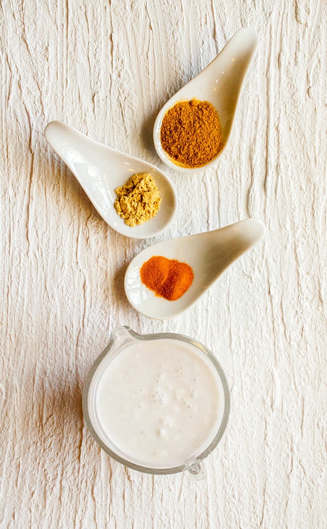 Pitcher of coconut milk with spoonfuls of turmeric, curry powder, and powdered ginger birds eye view.