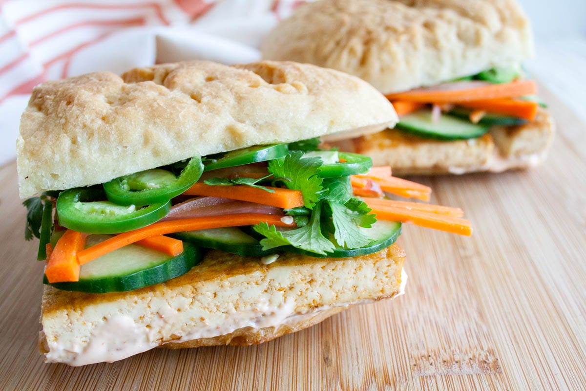 This Vietnamese Spicy Tofu Bánh Mì has spicy tofu, pickled carrots, sriracha mayo, cucumber, and jalapeño on a baguette. (vegan, gluten free) Tofu Bánh Mì on a cutting board.