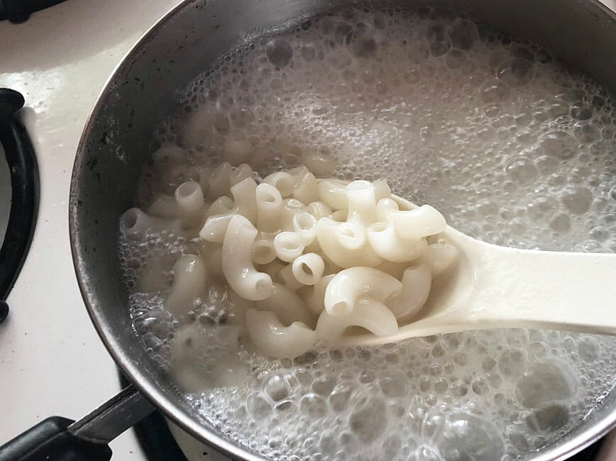 Cooking process of macaroni noodles in a large saucepan with salted water and spoon.