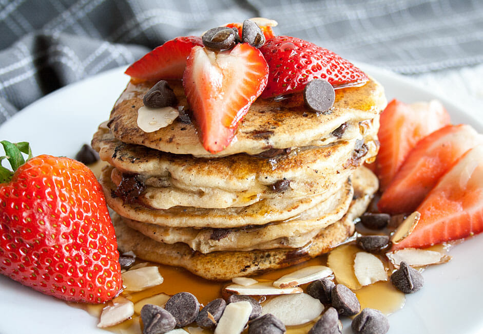 Fluffy Vegan Chocolate Chip Pancakes on a plate with strawberries, chocolate chips and a drizzle of maple syrup on top.