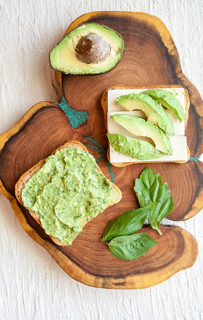 Slices of bread on a cutting board with avocado pesto, vegan mozzarella, and slices of avocado on top.