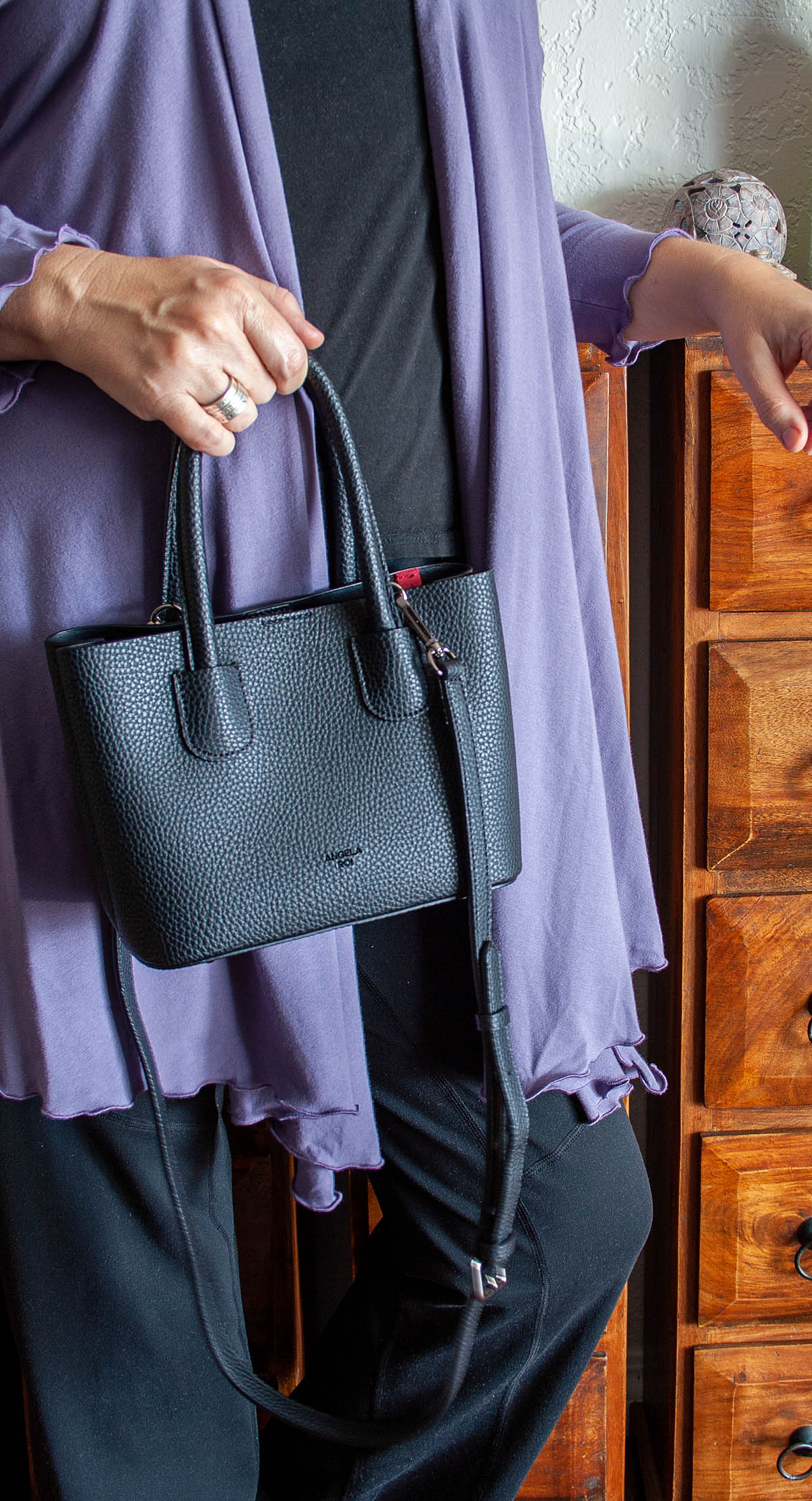 Photo of me in black pants, black t-shirt, lavender cover up and black Cher Micro handbag in my hand.