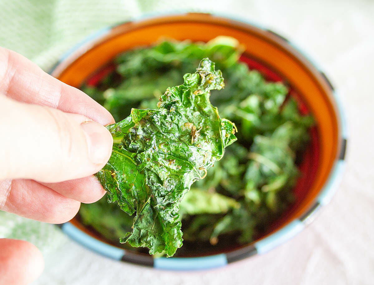 Roasted Ranch Kale leaf in hand with bowlful of kale chips in the background.