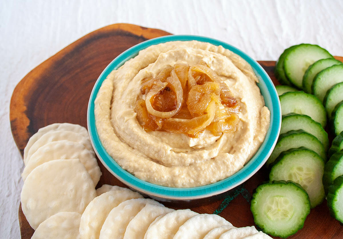 Caramelized Onion Hummus on cutting board with rice crackers and cucumber.