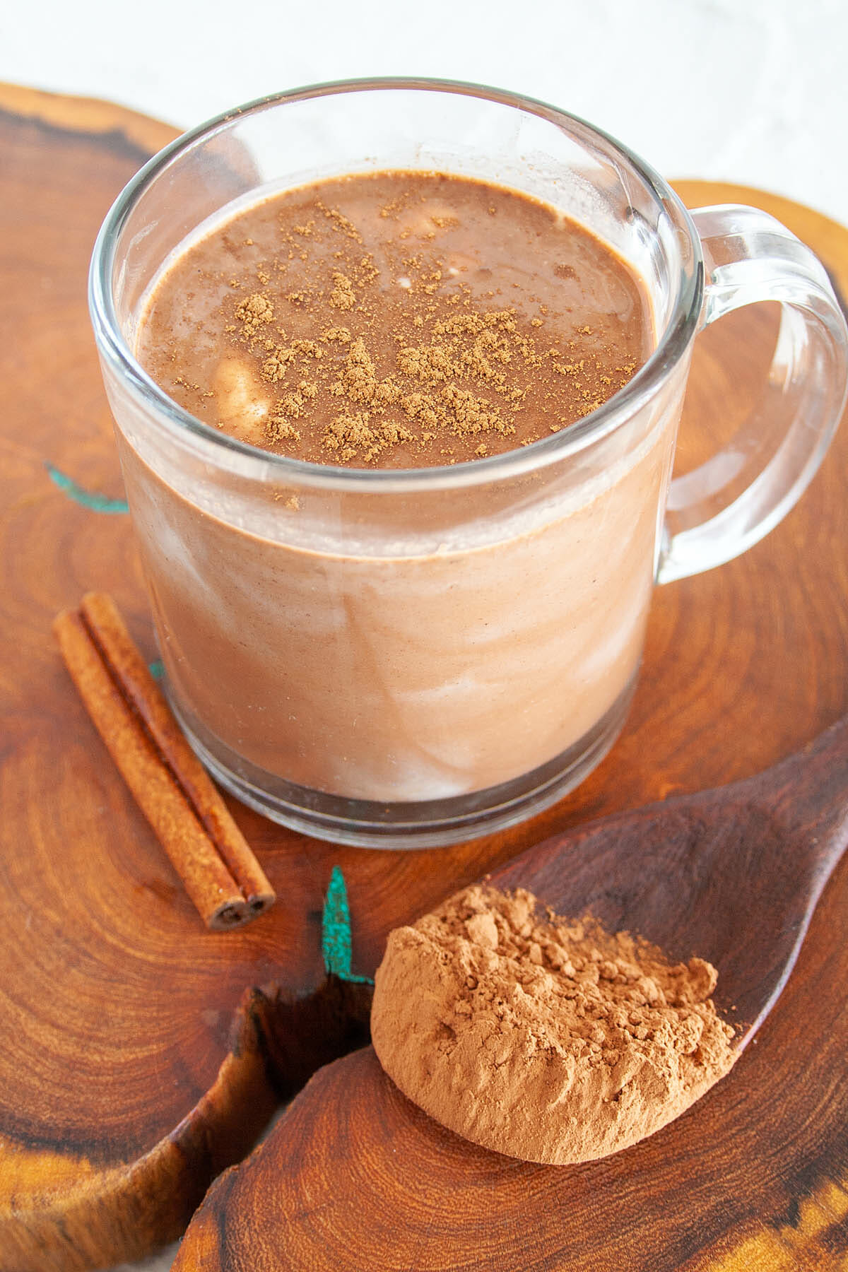 Spiced Hot Cocoa in mug on cutting board with spoonful of cacao powder and cinnamon stick.