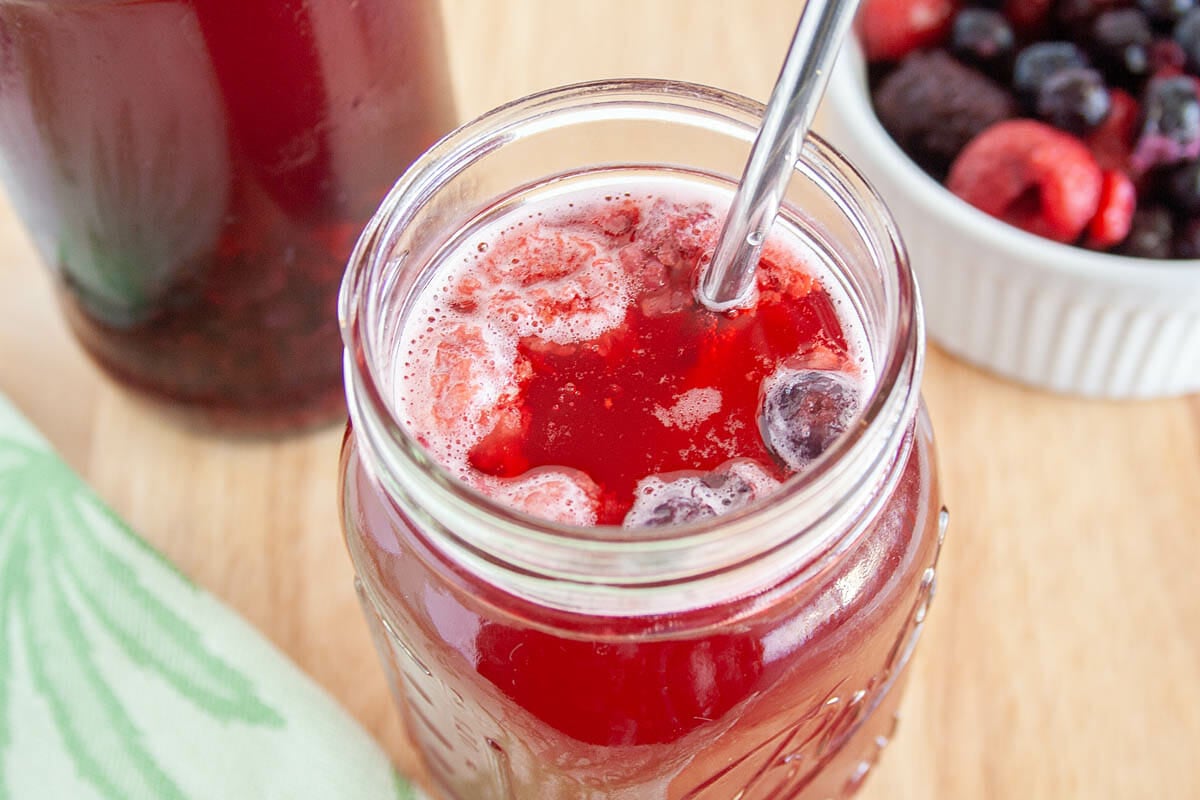 Mixed Berry Kombucha in a mason jar close up. Berries and bottle of kombucha in the background.