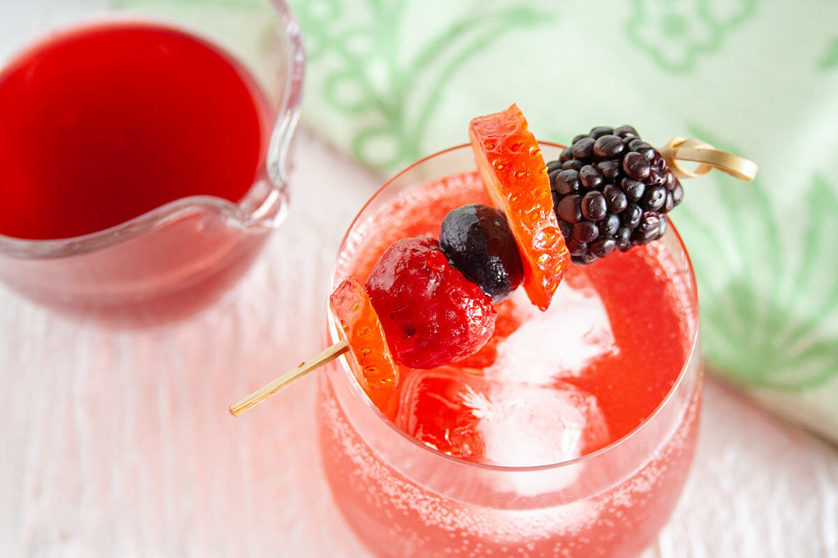 Mixed Berry Shrub mixed with club soda in a wine glass with ice cubes. Shrub in a pitcher in the background.