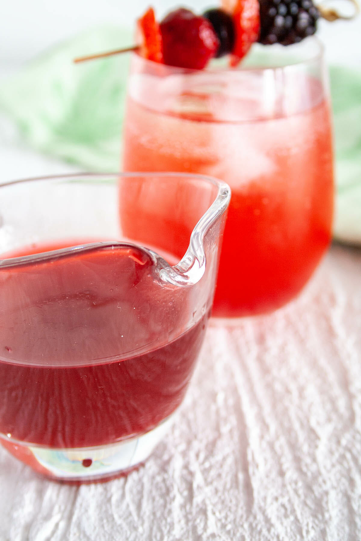 Mixed Berry Shrub in a pitcher and in a glass mixed with club soda in the background.