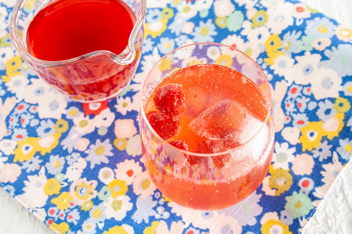 Raspberry Shrub in pitcher next to glass full of club soda and raspberry shrub.