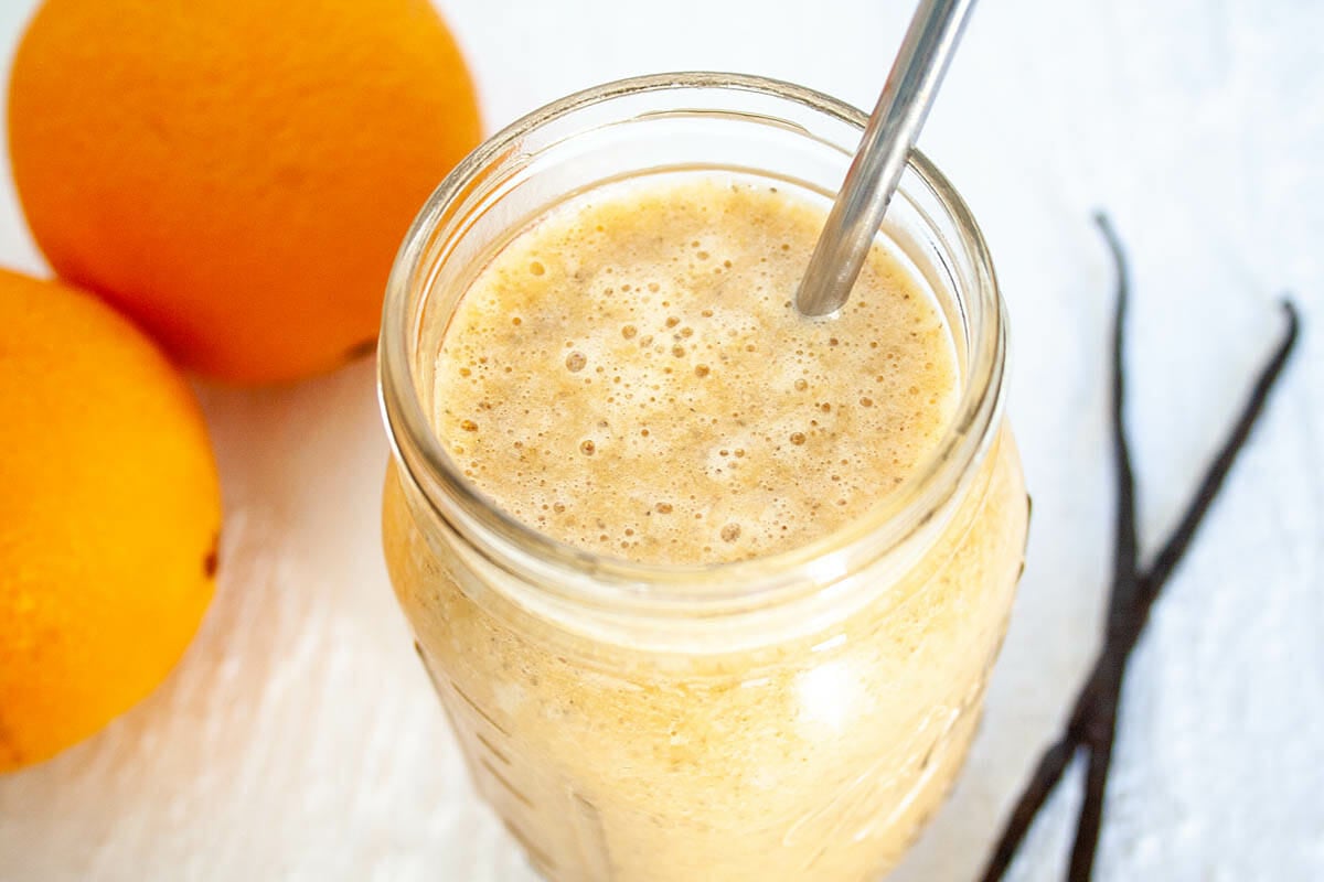 Vegan Orange Creamsicle Smoothie in mason jar with straw. There's oranges and vanilla bean in the background.
