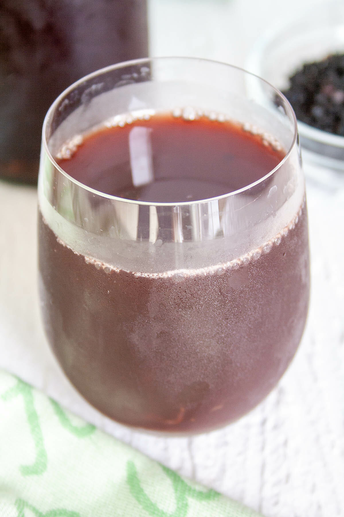 Elderberry Kombucha in a glass with bottle of kombucha and bowl of elderberries in the background.