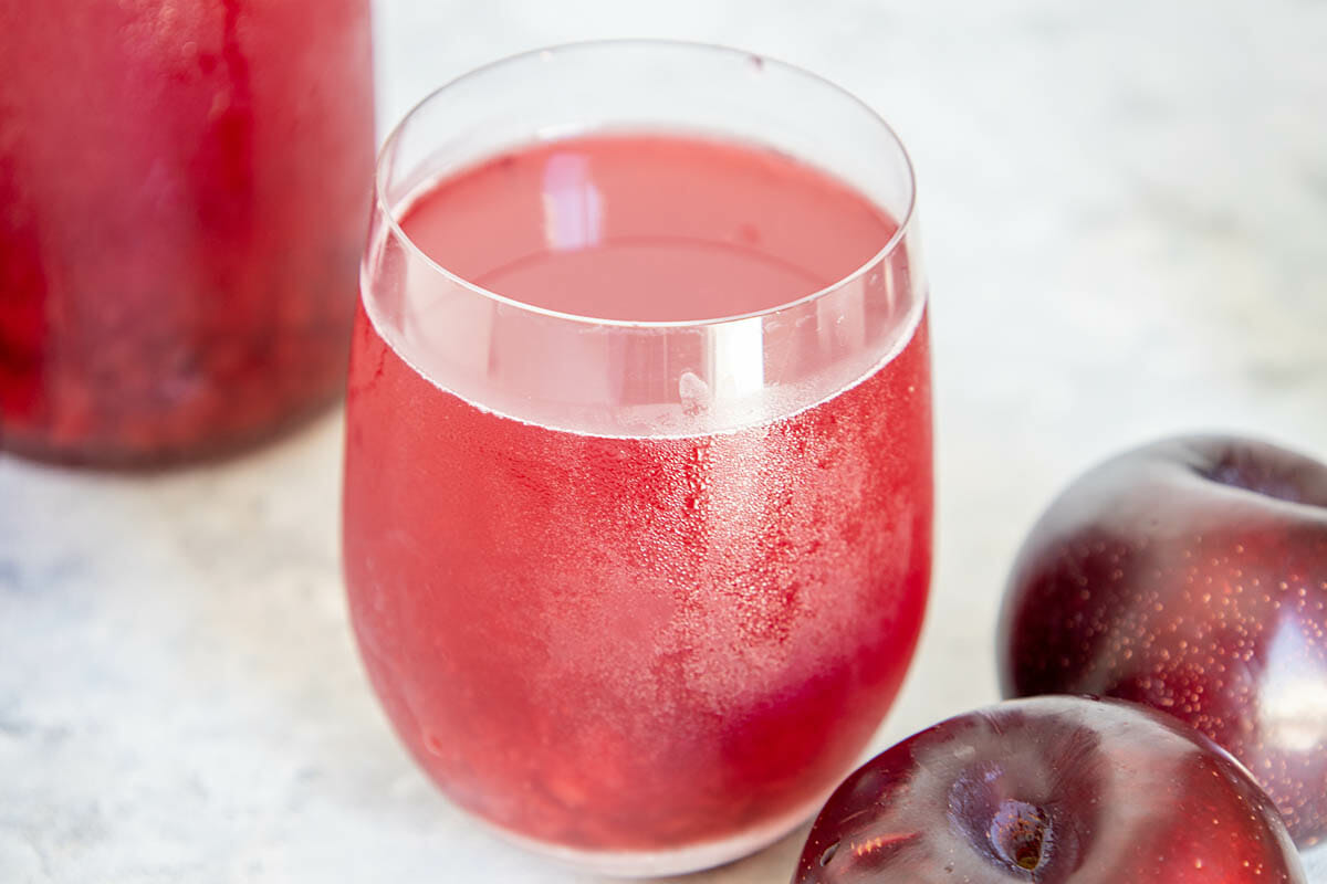 Plum Kombucha in a glass with plums and bottle of kombucha in the background.