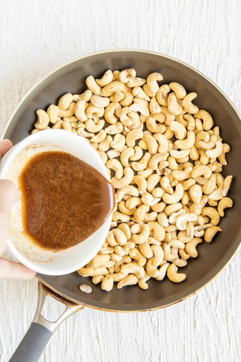 Cashews in a skillet with maple syrup mixture being added.