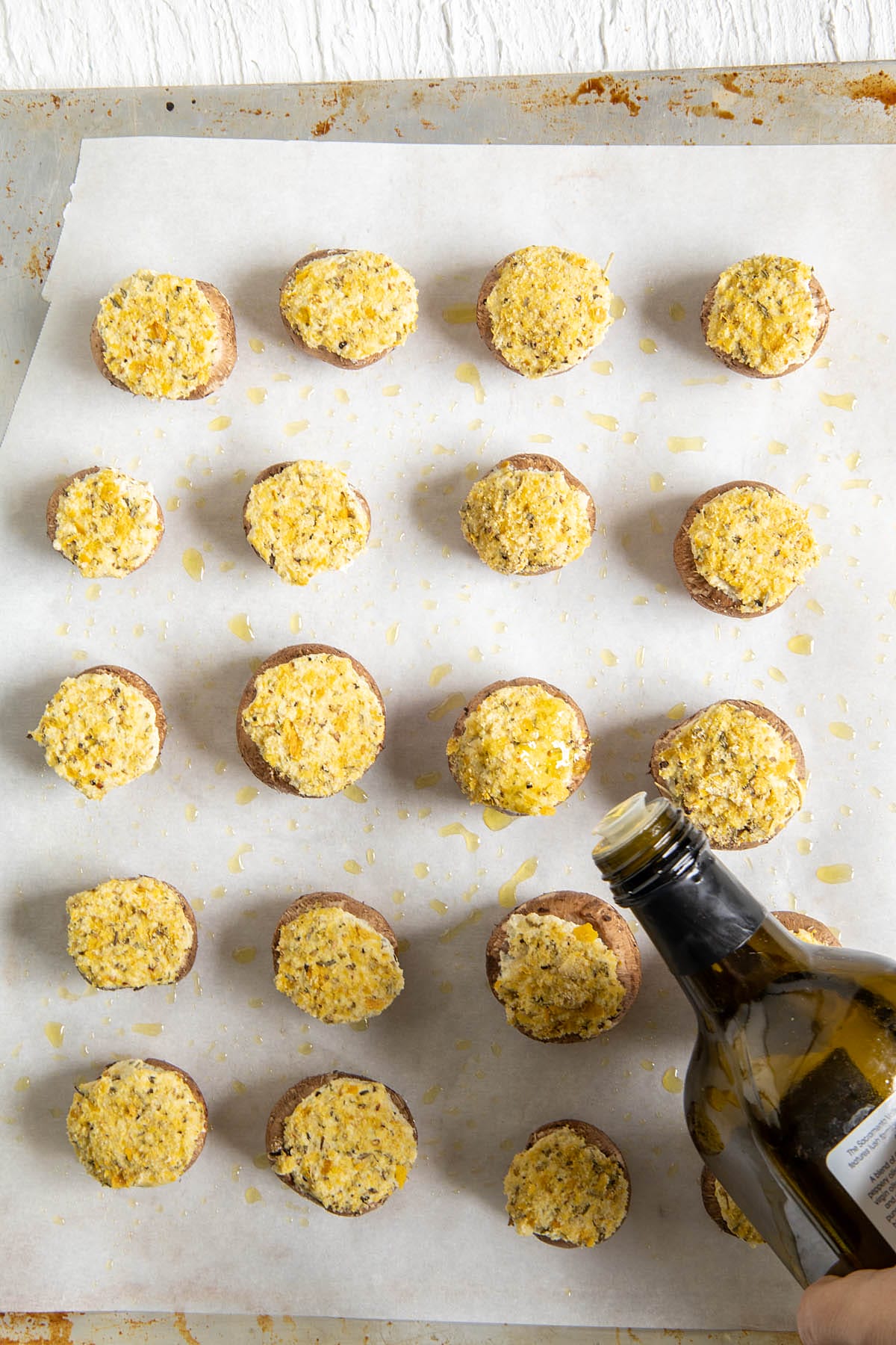 Olive oil being drizzled over stuffed and breaded mushrooms on a prepared baking sheet with oiled parchment paper.
