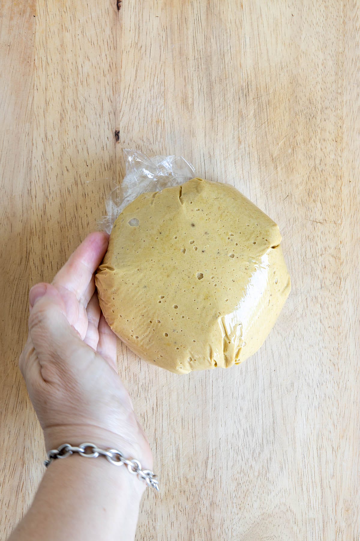 Cashew mixture wrapped in plastic wrap on a cutting board with hand shaping it.