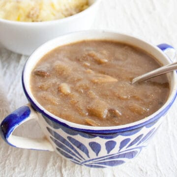 Vegan mushroom onion gravy in a bowl with spoon and mashed potatoes in the background.