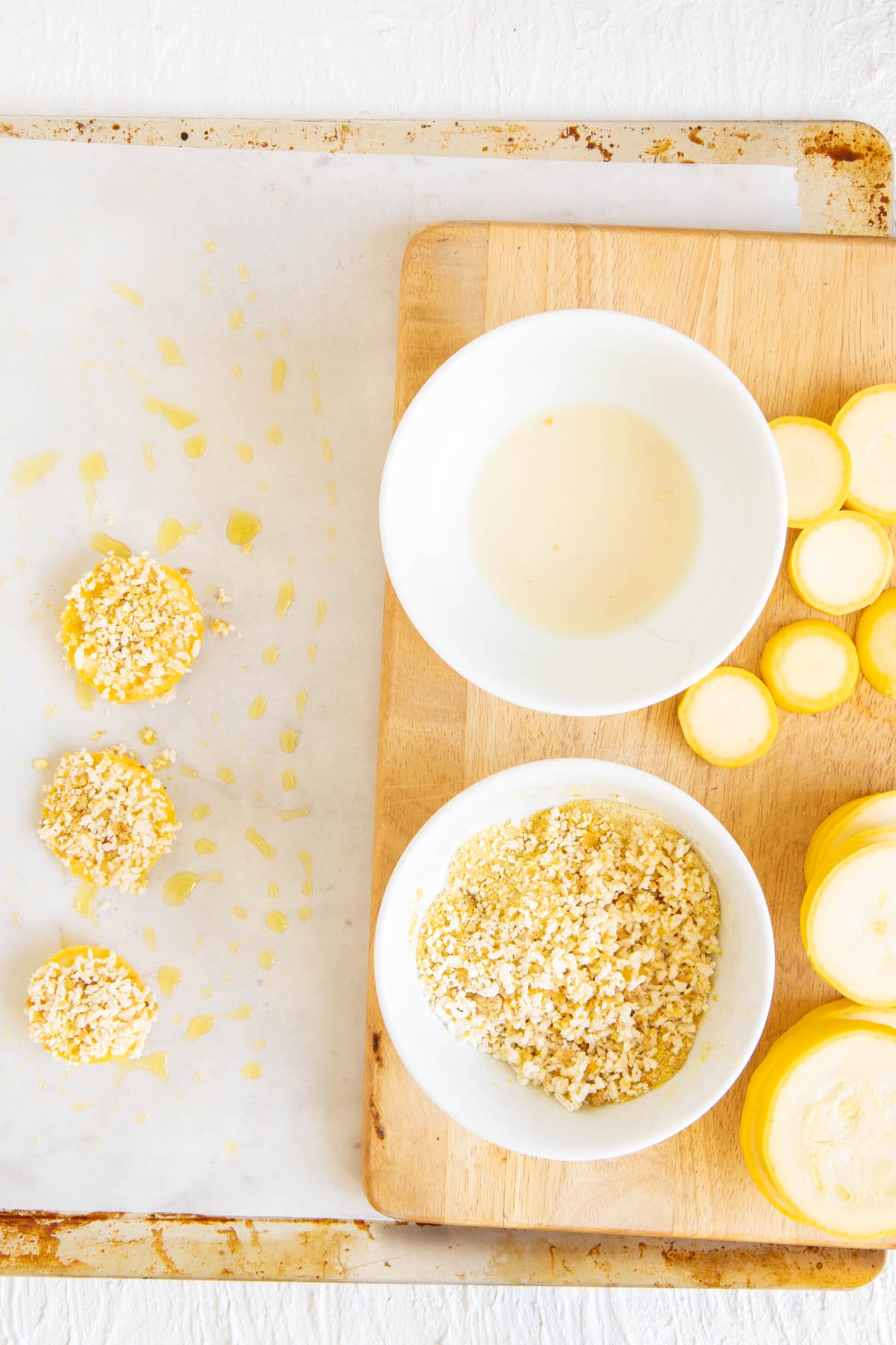 Squash slices with breading on parchment paper next to dipping station of bowlful of soy milk and bowlful of dry ingredients.