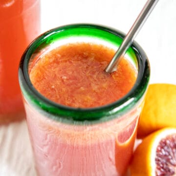 Blood Orange Kombucha in a glass with straw with a bottle of kombucha and blood oranges in the background.
