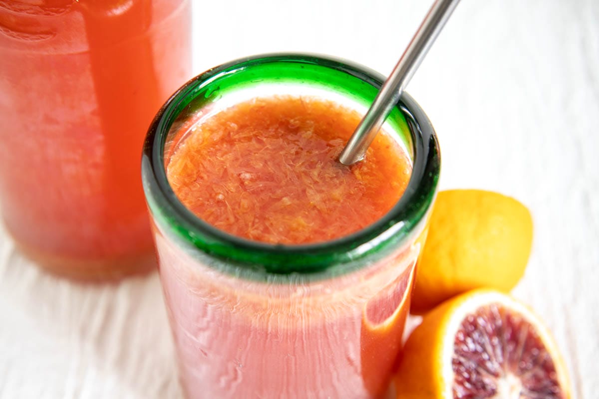 Blood Orange Kombucha in a glass with straw with a bottle of kombucha and blood oranges in the background.