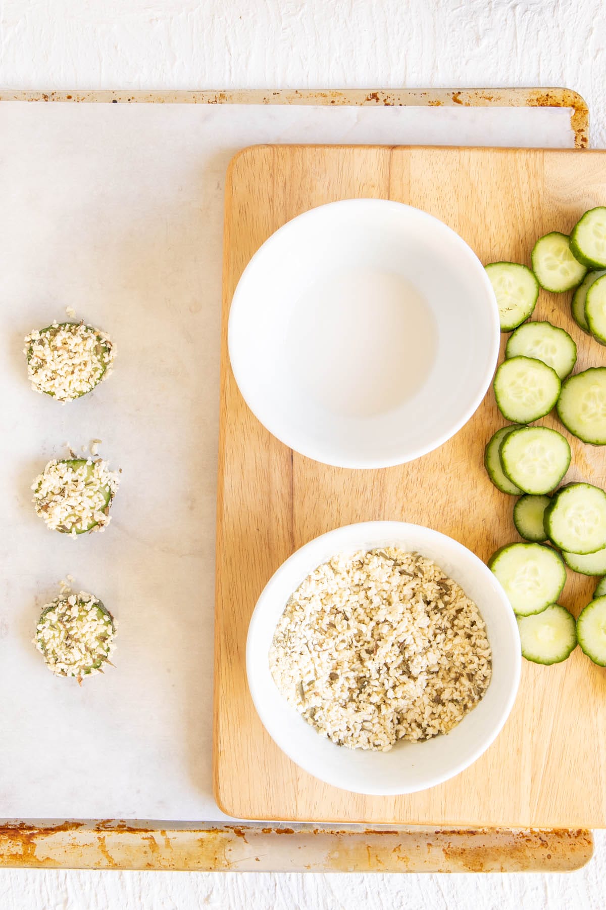 Sliced cucumber on a cutting board with bowl of dry mixture and bowl of soy milk. Battered cucumber on a parchment paper lined baking sheet.